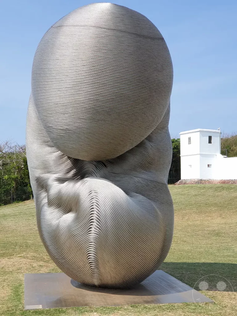 Eine massive, drahtgewickelte Skulptur dominiert den Vordergrund einer gepflegten Rasenfläche. Im Hintergrund erhebt sich das markante, weiße Gebäude des Eluanbi Leuchtturms unter einem strahlend blauen Himmel. Die Arbeit des Künstlers Kang Muxiang vermittelt ein Gefühl von dynamischer Form und beständiger Erneuerung, wie in "Sculpture Rebirth".