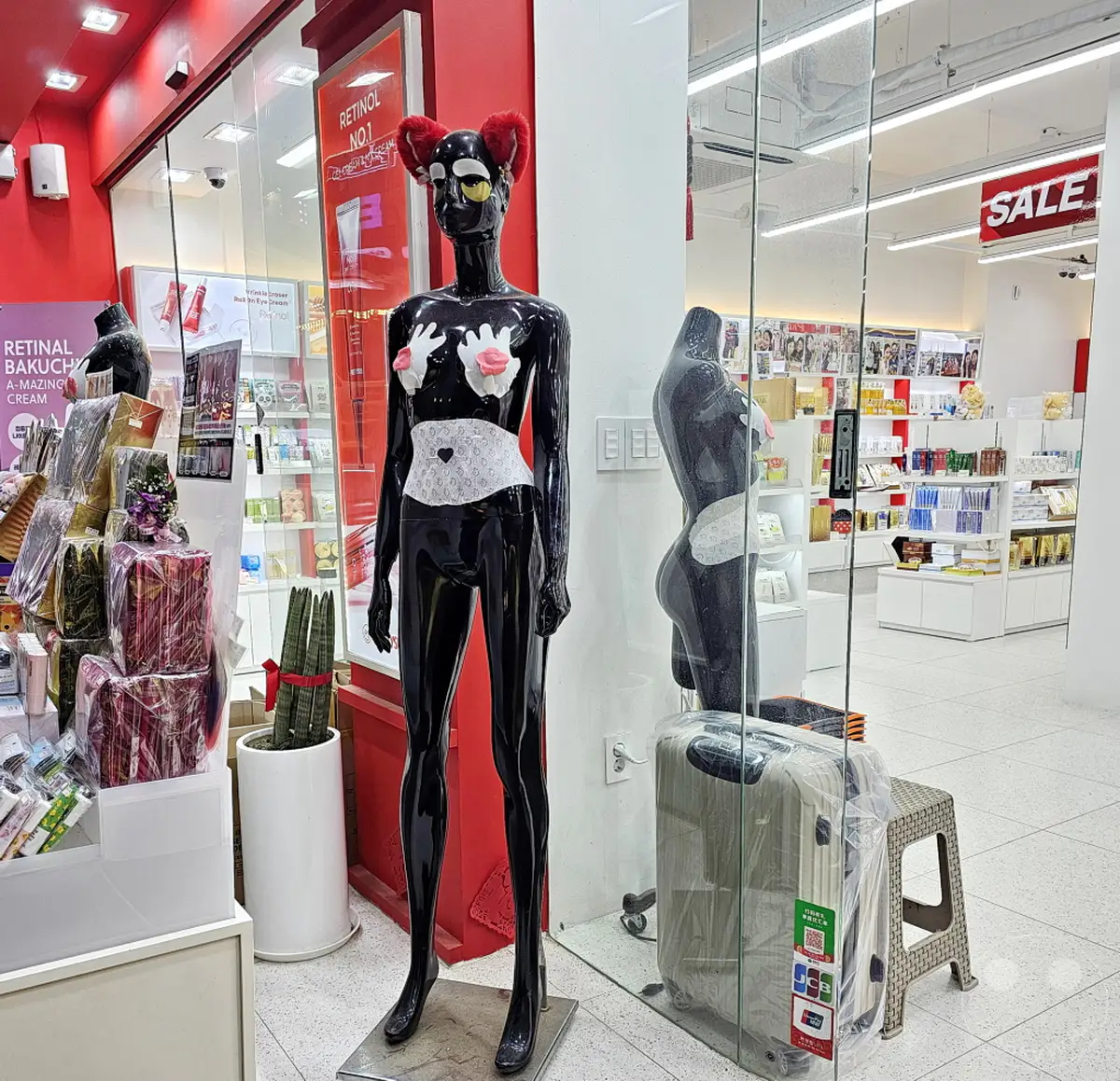 In Seoul, Südkorea, steht eine auffällige schwarze Schaufensterpuppe mit roten Fellohren und einer weißen Taillenband in einem Kosmetikgeschäft in Myeongdong Street. Im Hintergrund sind Regale mit Kosmetikprodukten und ein "Sale"-Schild zu sehen, was auf ein geschäftiges Einkaufsambiente hindeutet. Eine weitere, weniger detaillierte Schaufensterpuppe spiegelt sich im Glas neben einem Koffer.