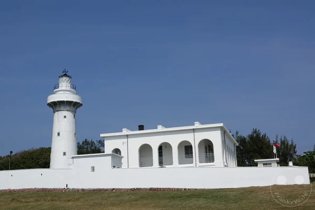 Der Eluanbi Lighthouse steht majestätisch vor einem strahlend blauen Himmel in Taiwan. Die strahlend weiße Architektur des Leuchtturms und des angrenzenden Gebäudes mit seinen Bögen bildet einen eindrucksvollen Kontrast zur grünen Wiese und den Bäumen im Hintergrund. Die Szene strahlt Ruhe und Beständigkeit aus, typisch für einen Küstenwachturm.