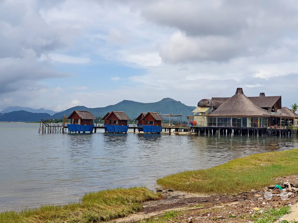 Die Lap An Lagune in Vietnam ist gesäumt von malerischen Fischerhütten auf Stelzen, die im sanften Wasser ruhen. Im Hintergrund erheben sich majestätische Berge unter einem dramatisch bewölkten Himmel, während ein großes, reetgedecktes Gebäude am Ufer zum Verweilen einlädt. Die Szene vermittelt eine friedliche, ländliche Atmosphäre, die durch die natürliche Schönheit der Umgebung geprägt ist.