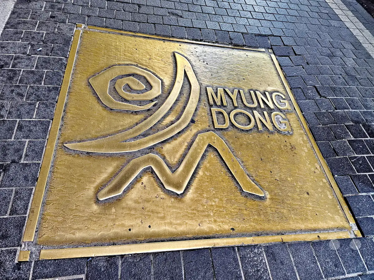 Eine auffällige Bronzeplakette mit dem Schriftzug "MYEONG DONG" und einem stilisierten Symbol ist in einen Gehweg aus dunklen Pflastersteinen eingelassen. Das strahlende Design auf dem Boden dient als Markierung für eine belebte Gegend in Seoul. Die Aufnahme erzeugt eine Atmosphäre von städtischer Erkundung und Entdeckung.
