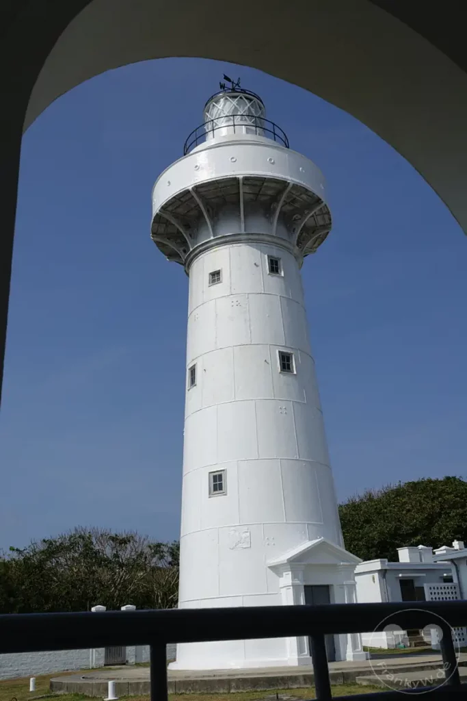 Der Eluanbi Lighthouse in Taiwan ragt majestätisch gegen einen strahlend blauen Himmel auf. Seine weiße, zylindrische Struktur mit zahlreichen kleinen Fenstern und einem markanten Dach dominiert die Szene, eingerahmt von einem architektonischen Bogen. Die umgebende Vegetation und ein angrenzendes Gebäude vervollständigen den Eindruck eines historischen und wichtigen maritimen Wahrzeichens.