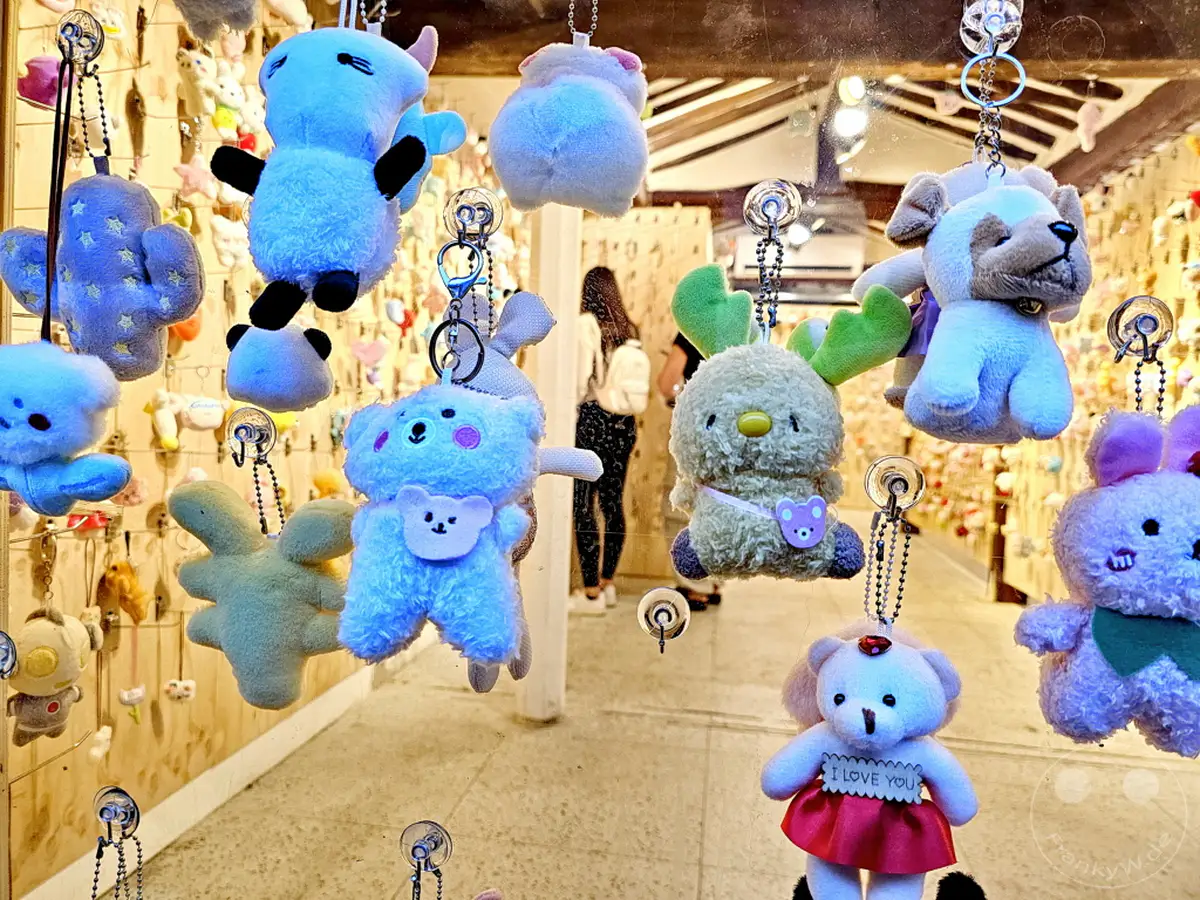 Zahlreiche süße Plüschtier-Schlüsselanhänger hängen an einer Fensterscheibe, darunter verschiedene Tiere wie Bären, Hunde und andere Fantasiewesen. Die Spielzeuge in Pastellfarben und mit kleinen Details strahlen eine verspielte und liebenswerte Atmosphäre aus, typisch für die charmanten Gassen des Ikseon-dong Hanok Village in Seoul. Der Hintergrund deutet auf einen traditionellen koreanischen Stil mit Holzelementen und weiteren Auslagen von Souvenirs hin.