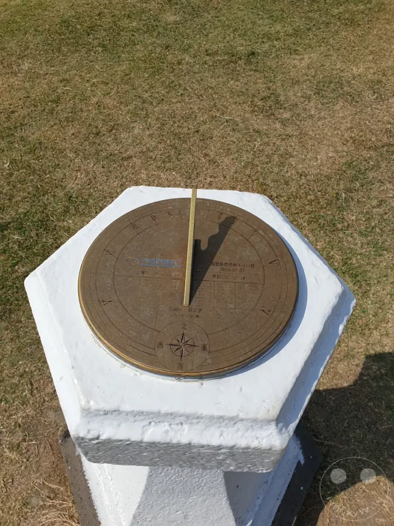 Eine historische Sonnenuhr am Eluanbi Lighthouse auf Taiwan zeigt die Mittagszeit an. Der Schatten des Gnomons wirft eine deutliche Linie über die bronzefarbene Zifferblatt, umgeben von grüner Graslandschaft. Die Szene strahlt eine ruhige und sonnige Atmosphäre aus, die zum Verweilen einlädt.