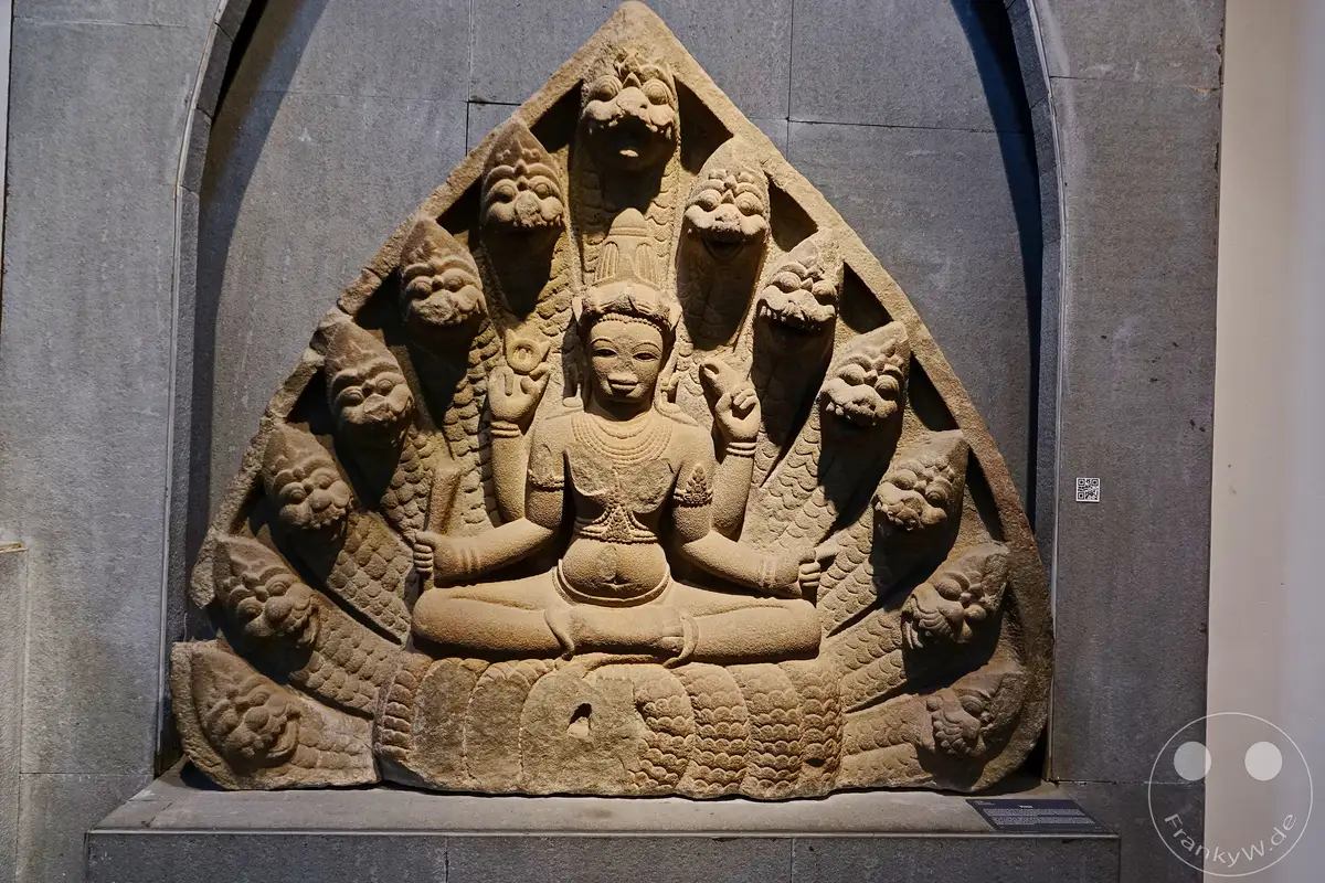 Eine steinerne Skulptur aus Vietnam, ausgestellt im Museum of Cham Sculpture in Da Nang, stellt eine zentrale Figur dar, die von sieben Naga-Schlangenköpfen umgeben ist. Die detailreiche Darstellung vermittelt eine Aura von archaischer Kunst und spiritueller Bedeutung. Die weiche Beleuchtung hebt die Textur des alten Steins und die kunstvollen Schnitzereien hervor.