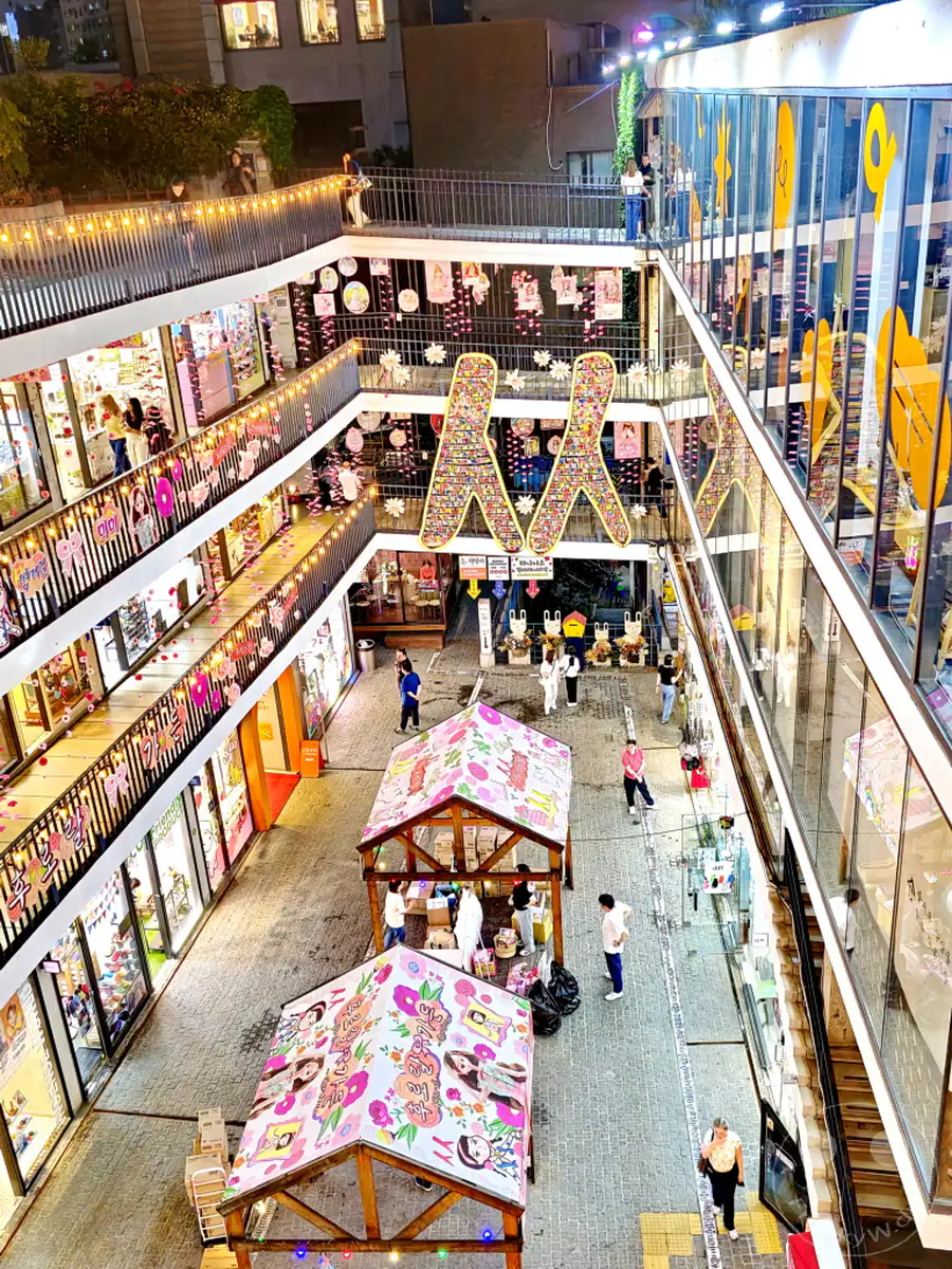 Blick von oben auf eine belebte Straße in Insadong, Seoul, Südkorea, die von Geschäften gesäumt ist. Kleine Marktstände mit bunten Überdachungen lockern das Bild auf, während Besucher zwischen den Gängen flanieren. Die festliche Beleuchtung und die lebhafte Atmosphäre laden zum Bummeln und Entdecken ein.