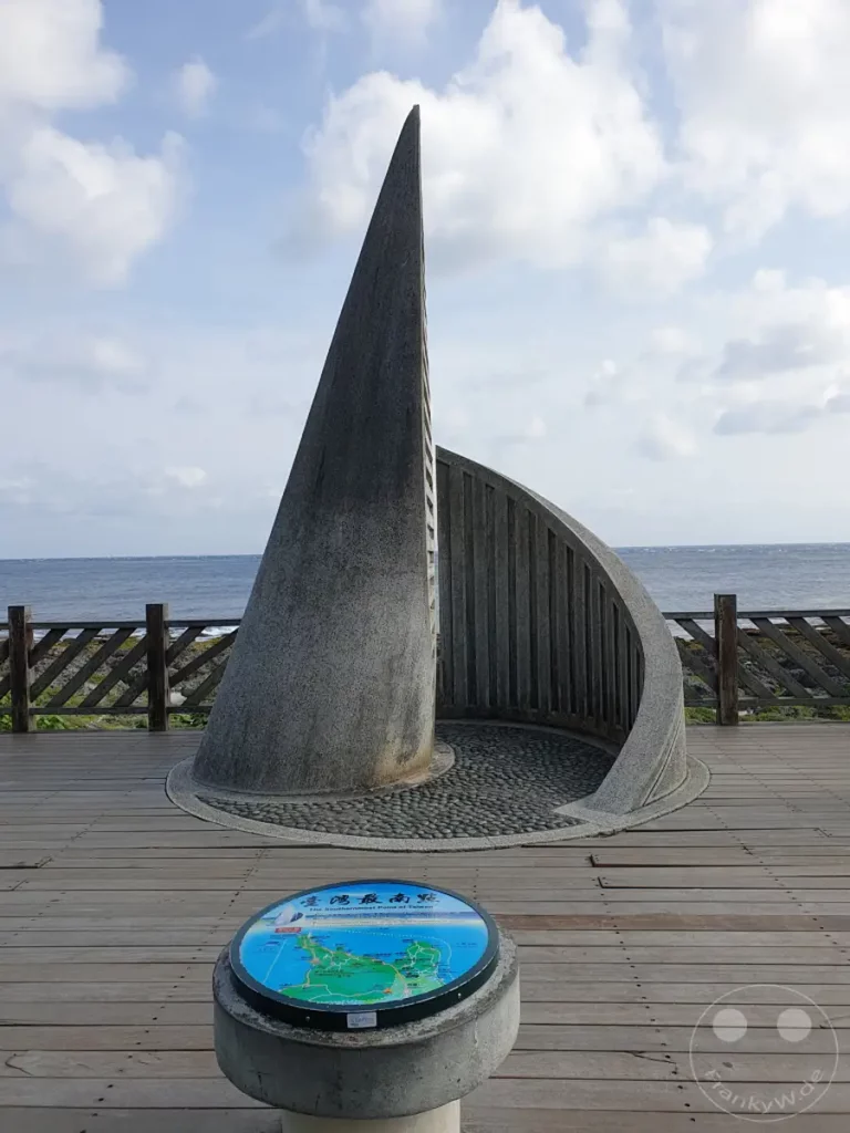 Das imposante Steindenkmal markiert den südlichsten Punkt Taiwans und bietet einen atemberaubenden Blick auf das Meer. Kleine Kieselsteine bedecken den Boden innerhalb des Denkmals, während Holzplanken zu einer Aussichtsplattform führen. Wolken ziehen über den blauen Himmel und unterstreichen die friedliche Atmosphäre dieses Küstenabschnitts.