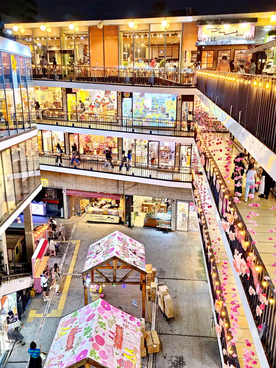 Eine belebte Nachtszene in Insadong Street, Seoul, Südkorea, zeigt einen Marktplatz mit mehreren Ebenen voller Geschäfte und Stände. Lichterketten und rosa Dekorationen erhellen die Gänge und schaffen eine festliche Atmosphäre, während Besucher zwischen den Läden schlendern. Der Blick von oben enthüllt die lebendige und geschäftige Energie des Viertels bei Nacht.