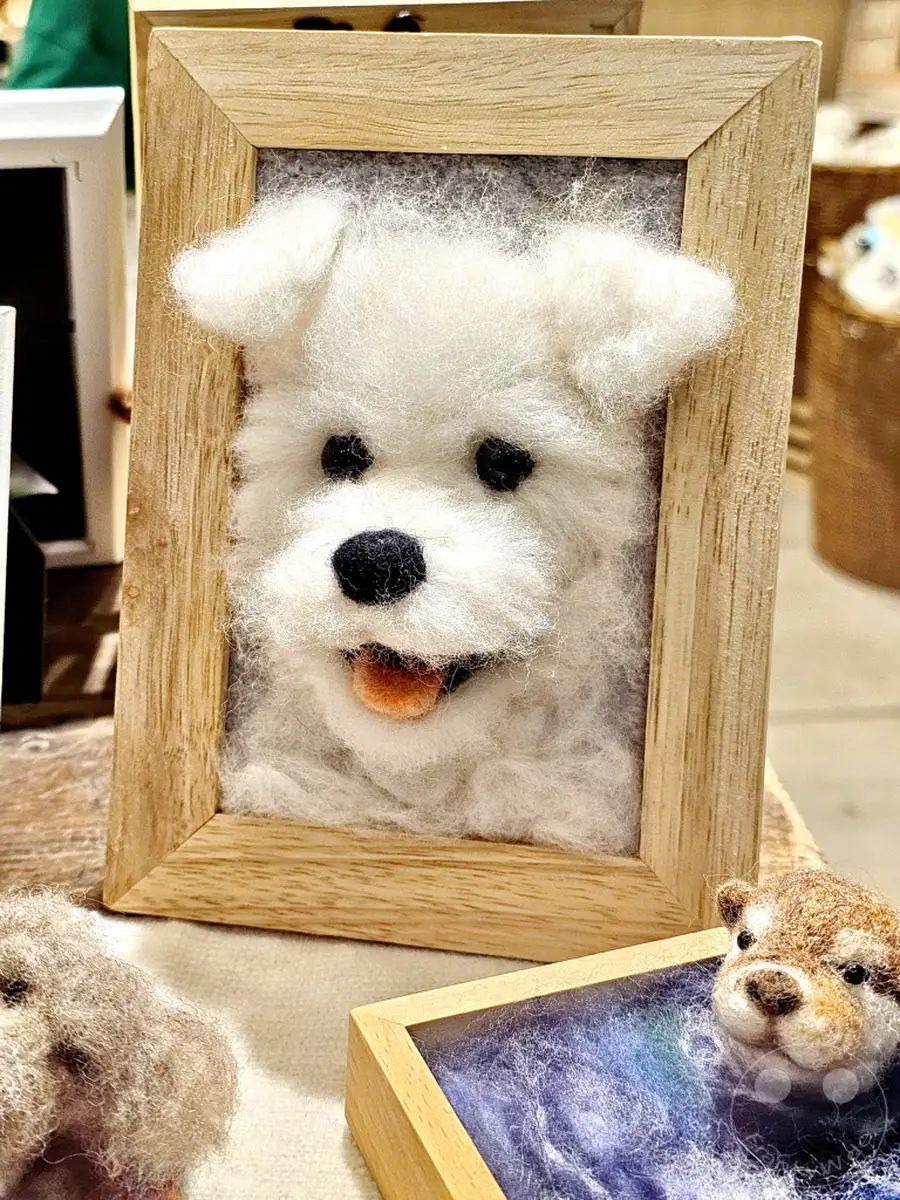 Ein detailreiches, handgefertigtes Porträt eines weißen Hundes in einem Holzrahmen steht im Mittelpunkt des Bildes in Insadong, Seoul, Südkorea. Das Kunsthandwerk strahlt eine Wärme und Niedlichkeit aus, die typisch für die kreative Atmosphäre der Insadong Street ist. Im Vordergrund sind weitere kleine, filzige Tierfiguren zu sehen, die das charmante und künstlerische Ambiente des Ortes unterstreichen.