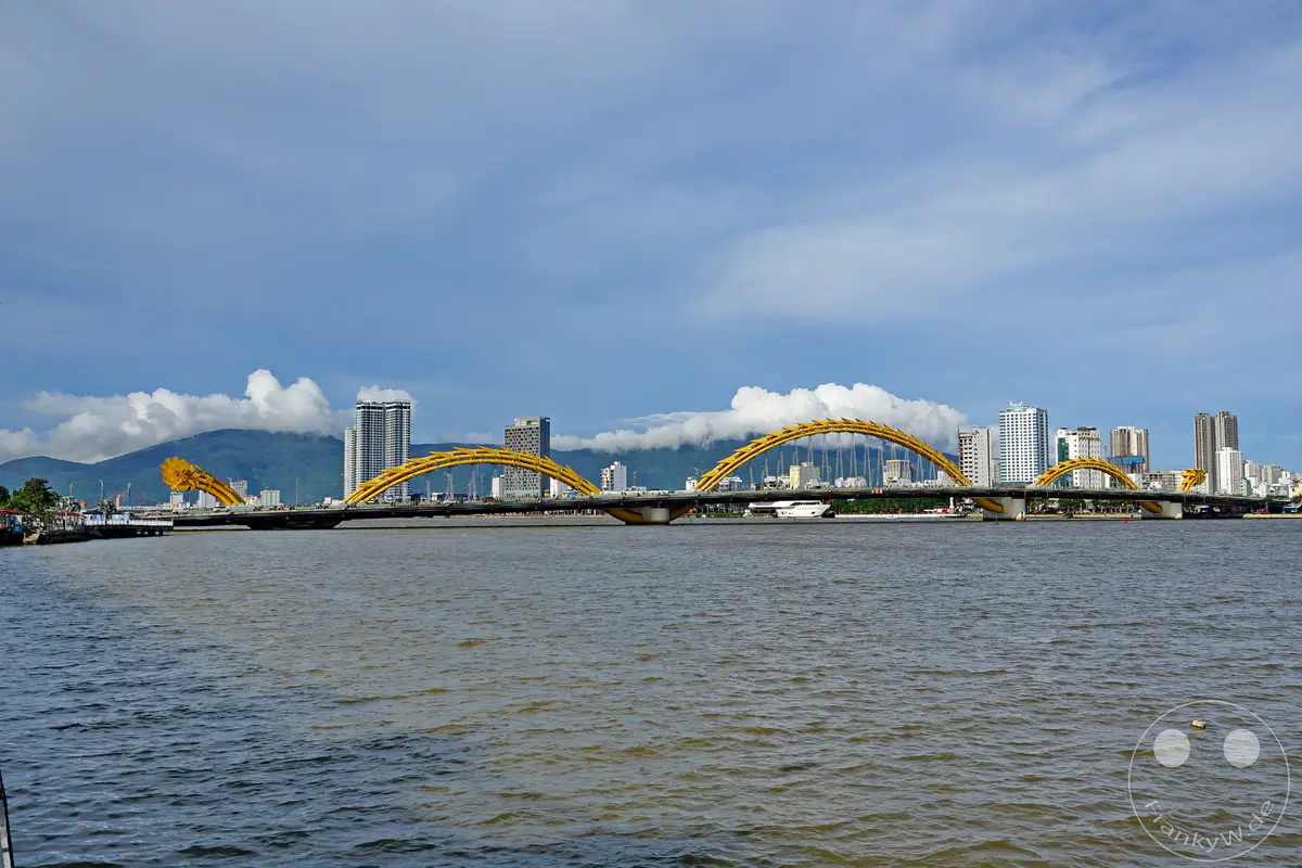 Die markante Dragon Bridge überspannt in Da Nang, Vietnam, den Han-Fluss und symbolisiert die asiatische Kultur. Hohe moderne Gebäude säumen das Ufer und bilden eine beeindruckende Skyline im Hintergrund. Das ruhige Wasser des Flusses reflektiert die Architektur und schafft eine harmonische Atmosphäre unter dem weiten Himmel.