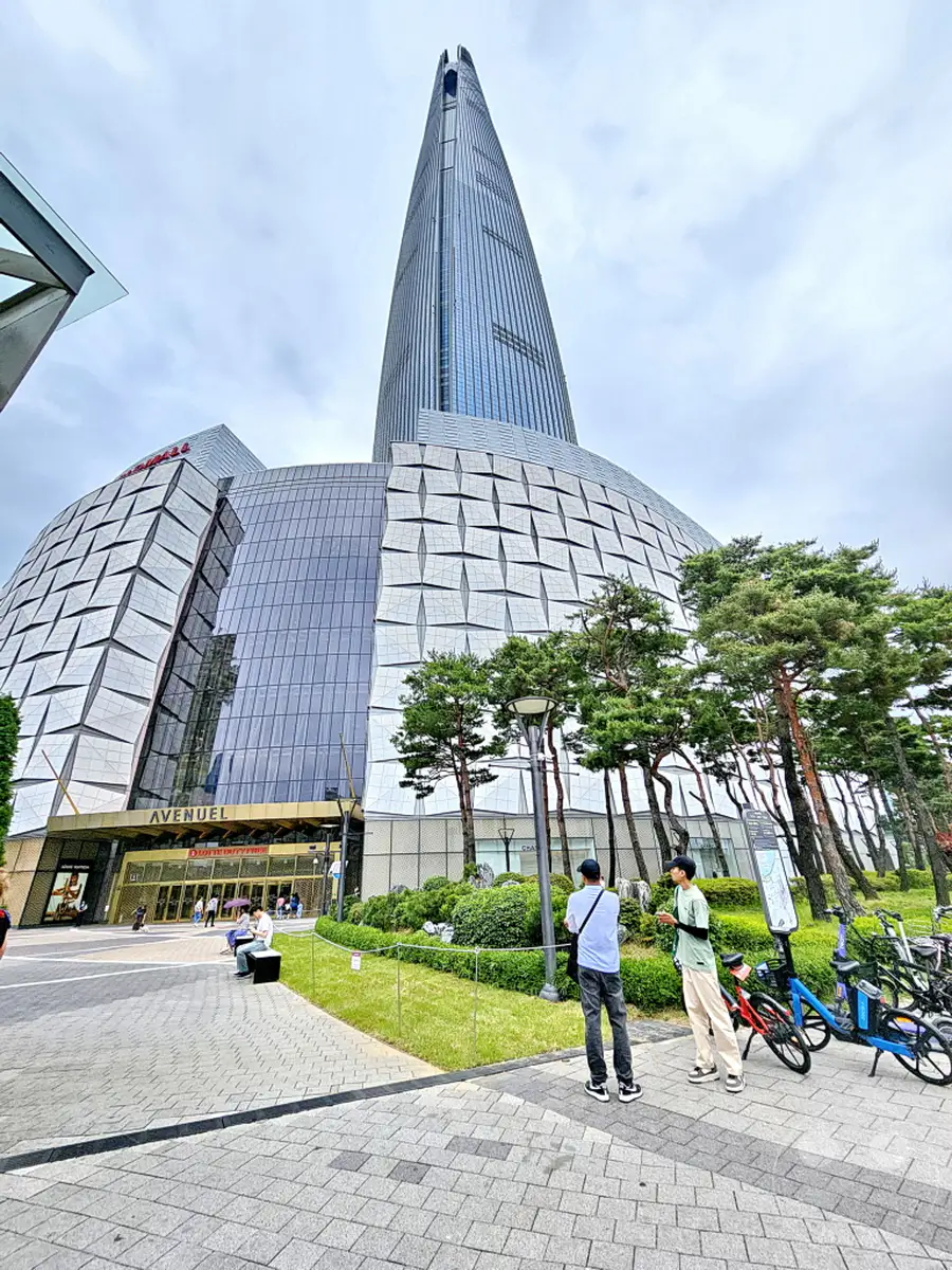 Südkorea - Seoul - Der imposante Lotte World Tower überragt die futuristische Architektur des angrenzenden Einkaufszentrums. Vor dem Eingangsbereich des Avenuel sind einige Bäume und Büsche angelegt, die eine grüne Oase inmitten der urbanen Kulisse bilden. Zwei Männer stehen im Vordergrund und unterhalten sich, während im Hintergrund Fahrräder zum Verleih bereitstehen.