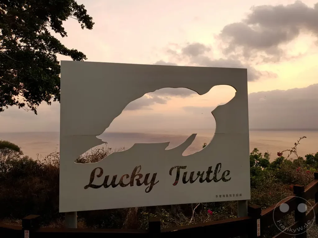 Ein kreatives Schild mit einer stilisierten Schildkröte steht im Vordergrund und bietet durch seine Silhouette einen Blick auf das Meer bei Sonnenuntergang. Die warme Abendsonne taucht den Himmel und das Meer in sanfte Farben, während Wolken dramatische Formen bilden. Die Szenerie vermittelt eine friedliche und malerische Atmosphäre an der Küste Taiwans, wahrscheinlich in der Nähe von Guanshan (Mt. Guan).
