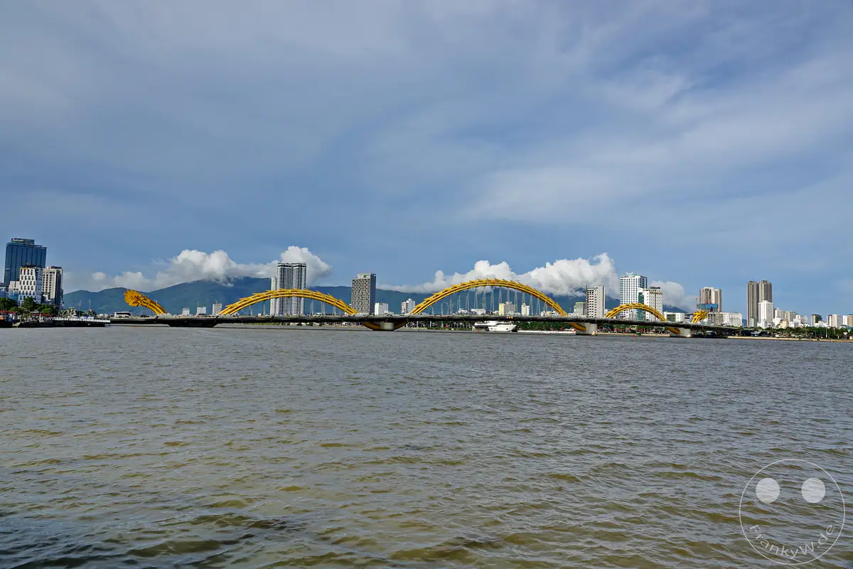 Die Drachenbrücke in Da Nang, Vietnam, überspannt den Han-Fluss und ist ein markantes Wahrzeichen der Stadt. Moderne Hochhäuser säumen das Ufer und bilden einen Kontrast zur imposanten Brückenkonstruktion. Der Himmel ist von Wolken durchzogen, was der Szene eine leicht dramatische, aber dennoch ruhige Atmosphäre verleiht.