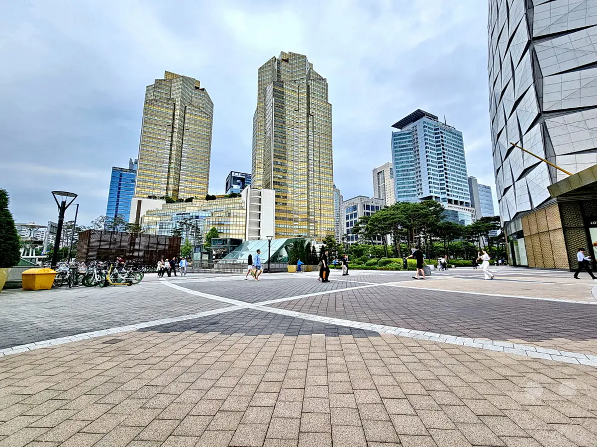 Südkorea, Seoul – Eine weitläufige Plaza mit modernen Wolkenkratzern im Hintergrund, darunter der Lotte World Tower. Menschen spazieren entspannt auf dem gepflasterten Platz, während Fahrräder am Rand geparkt sind. Die Szenerie ist geprägt von urbaner Architektur und einem bewölkten Himmel, was der Atmosphäre eine ruhige Dimension verleiht.