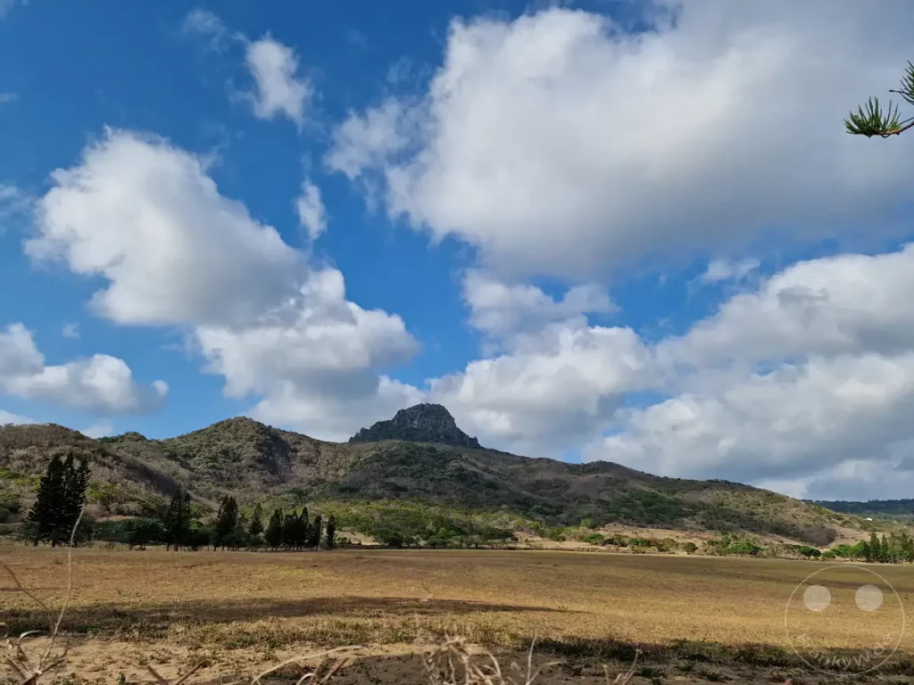 Eine weite, trockene Ebene erstreckt sich vor einer sanft ansteigenden Bergkulisse unter einem dramatischen Himmel mit großen, weißen Wolken. Grüne Bäume säumen den Fuß der Hügel, die von der Sonne ausgetrocknet wirken und eine leichte Melancholie ausstrahlen. Die Szene in Kenting, Taiwan, lädt zum Innehalten und zur Betrachtung der Natur ein.