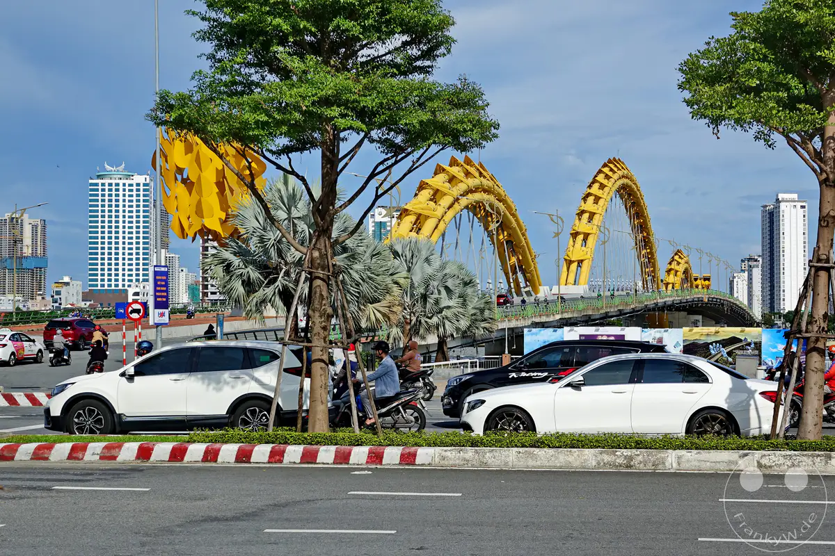 Die beeindruckende Dragon Bridge in Da Nang, Vietnam, überspannt den Fluss und bildet eine markante Silhouette in der Skyline der Stadt. Moderne Hochhäuser und geschäftiger Verkehr mit Autos und Motorrädern prägen das urbane Szenario. Die goldene Drachenbrücke ist ein Wahrzeichen, das die dynamische Atmosphäre der Stadt einfängt.