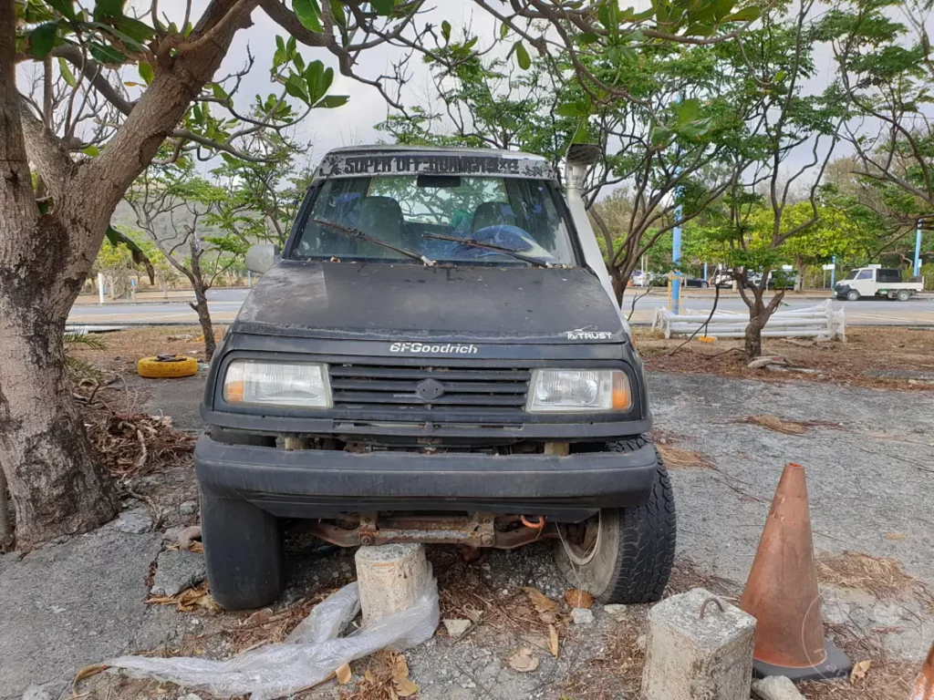Ein verwitterter Suzuki Vitara steht abseits einer Straße in Kenting, Taiwan, unter Bäumen. Der Geländewagen mit 'Super Off-Road'-Aufkleber und fehlender Stoßstange wirkt seitlich durch Betonblöcke abgestützt. Die Szene vermittelt eine Atmosphäre der Verlassenheit und des Stillstands inmitten natürlicher Vegetation.
