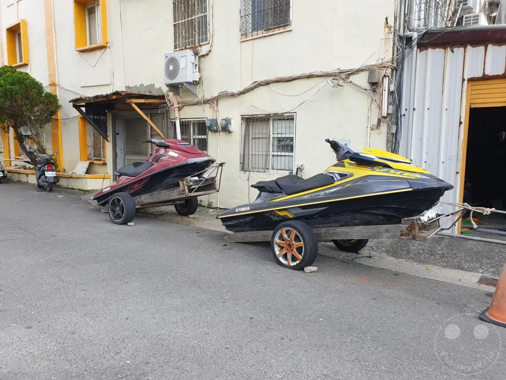 Zwei Jet-Skis auf Anhängern stehen auf einer asphaltierten Straße in Kenting, Taiwan. Ein roter Jet-Ski steht auf einem zweirädrigen Anhänger vor einem gelblich-weißen Gebäude, während ein gelb-schwarzer Jet-Ski auf einem ähnlichen Anhänger daneben positioniert ist. Die Szene vermittelt eine ruhige und alltägliche Atmosphäre, als ob die Wasserfahrzeuge gerade transportiert oder abgestellt wurden.