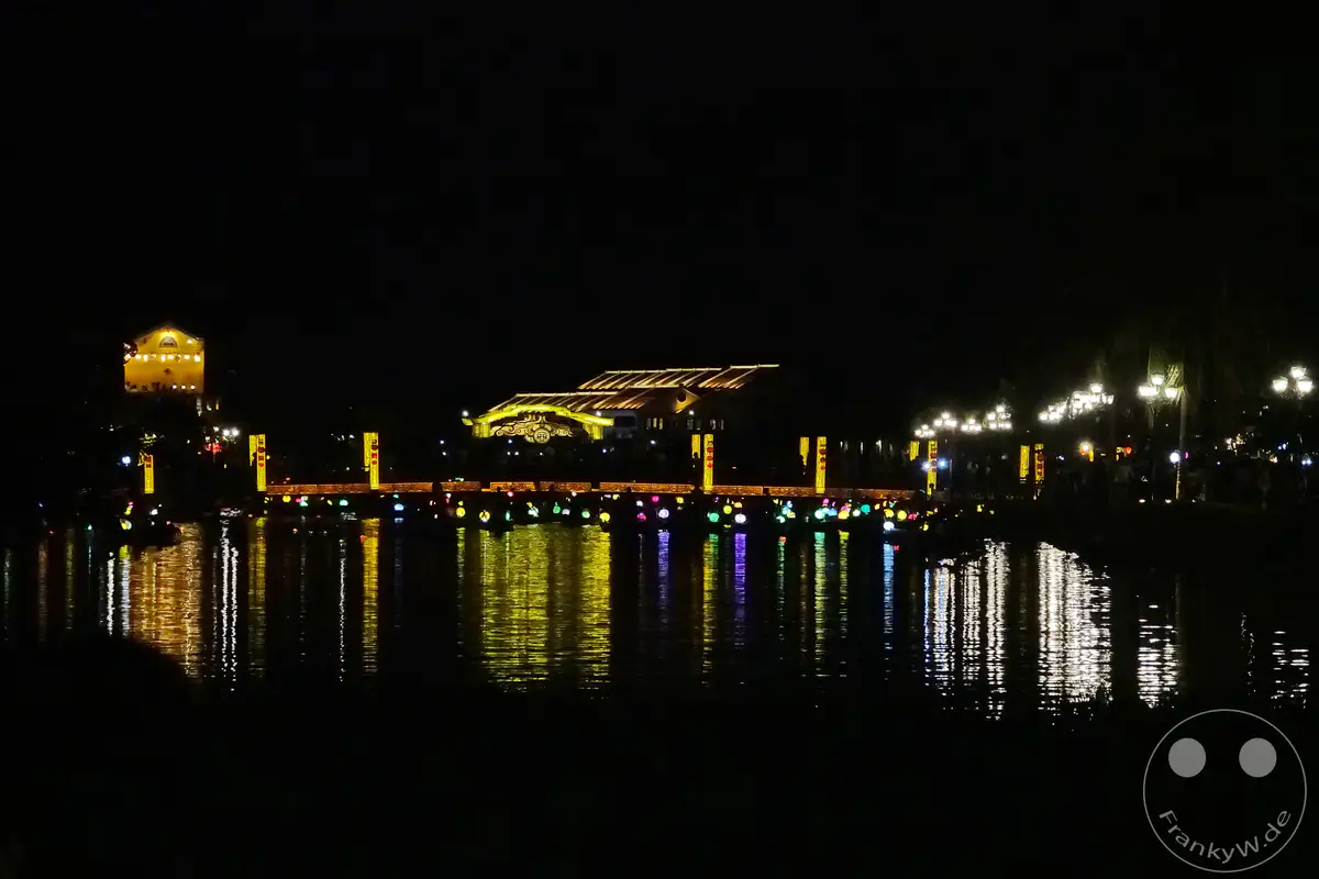 Eine beleuchtete Brücke überspannt nachts einen ruhigen Fluss in Hoi An, Vietnam. Bunte Lichter von Laternen spiegeln sich auf der Wasseroberfläche und erzeugen eine magische Atmosphäre. Die stimmungsvolle Nachtszene lädt zum Verweilen ein und fängt die romantische Ausstrahlung der Stadt ein.