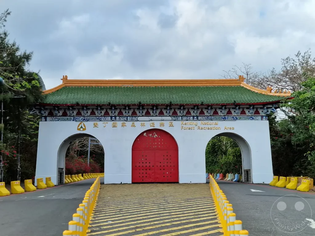 Der imposante Eingangsbogen des Kenting National Forest Recreation Area in Taiwan kündigt eine naturnahe Erkundung an. Die traditionelle asiatische Architektur mit dem grünen Ziegeldach und den roten Toren bildet einen farbenfrohen Kontrast zur umliegenden grünen Vegetation und dem bewölkten Himmel. Gelbe Absperrungen und Fahrbahnmarkierungen leiten Besucher auf dem Weg in den Erholungsbereich.