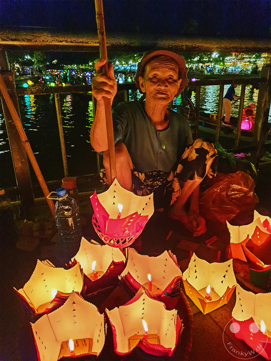 Eine ältere Frau sitzt nachts am Flussufer in Hoi An und verkauft schwimmende Laternen mit Kerzen. Im Hintergrund ist ein belebter Fluss zu sehen, auf dem Boote mit bunten Lichtern fahren und die festliche Atmosphäre der Nacht unterstreichen. Die Szene strahlt eine ruhige und magische Stimmung aus, typisch für das nächtliche Hoi An.