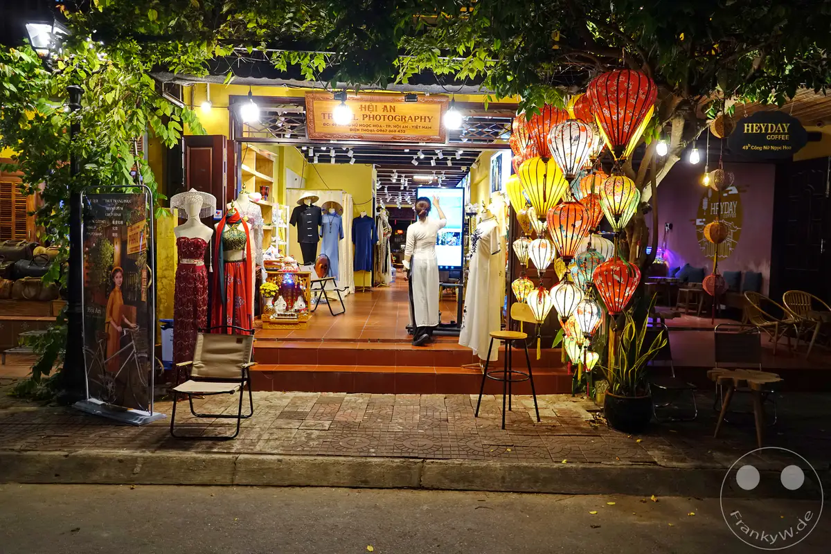 Eine Straße in Hoi An, Vietnam, wird bei Nacht durch bunte Lampions und die Auslagen eines Fotostudios beleuchtet. An der Fassade des Studios hängen traditionelle Kleider und Accessoires, während daneben Sitzgelegenheiten eines Cafés zum Verweilen einladen. Die Atmosphäre ist stimmungsvoll und lädt zum Flanieren und Entdecken ein.