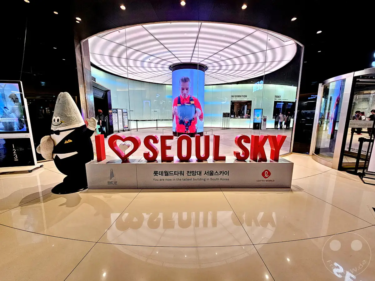 Ein freundlicher Star-Wars-inspirierter Charakter steht neben einer "I ❤️ SEOUL SKY"-Installation im Lotte World Tower. Die Installation wird von einem zylindrischen Bildschirm mit einem Bild eines Mannes eingerahmt, der etwas hält, und im Hintergrund ist die moderne Architektur des Gebäudes sichtbar. Die Szene vermittelt eine einladende und moderne Atmosphäre für Besucher in Seoul, Südkorea.