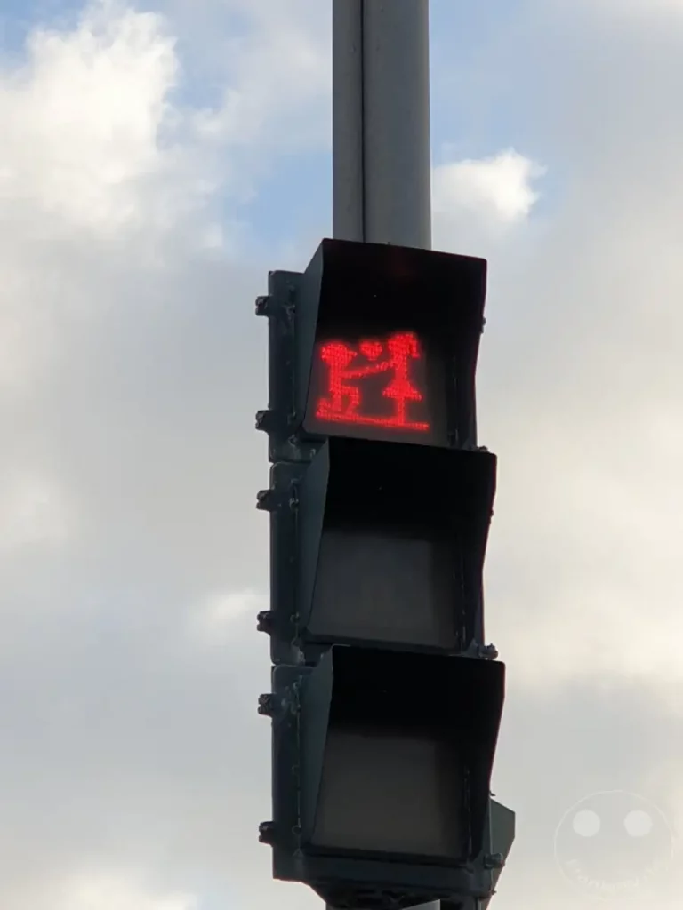 In Kenting, Taiwan, leuchtet eine rote Fußgängerampel mit einer besonderen Darstellung. Zwei stilisierte Figuren, die ein Paar darstellen, warten auf das Grünlicht. Die Szene wird durch den bewölkten Himmel und die klare Darstellung der Ampelsymbole geprägt.