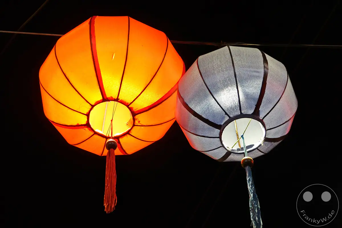 Zwei leuchtende Laternen, eine in leuchtendem Orange und die andere in hellem Weiß, hängen im Dunkeln und erhellen die Nacht von Hoi An, Vietnam. Die traditionellen Laternen mit ihren Stoffbehängen und kleinen Kugelelementen erzeugen eine festliche und magische Atmosphäre. Die Szene strahlt eine ruhige, aber lebendige Stimmung aus, typisch für eine Nacht in dieser historischen Stadt.
