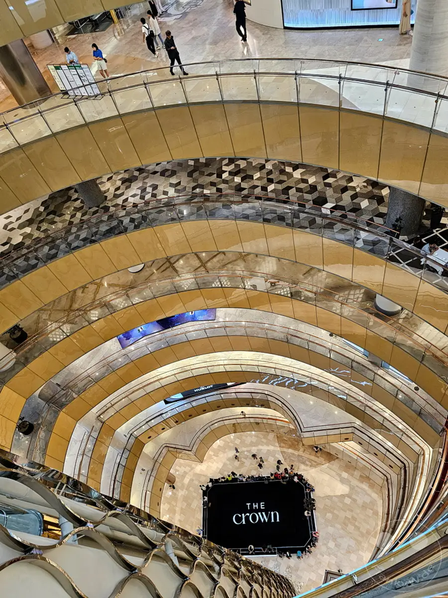 Blick von oben in das Atrium eines modernen Einkaufszentrums in Seoul, Südkorea. Die spiralförmige Architektur und die geschwungenen Ebenen des Lotte World Tower schaffen eine dynamische und beeindruckende Tiefenwirkung. Menschenmengen versammeln sich auf der unteren Ebene, um eine Veranstaltung vor einer großen schwarzen Leinwand zu verfolgen.
