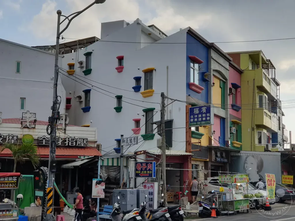 Eine bunte Straße in Kenting, Taiwan, mit farbenfrohen Gebäuden und Geschäften. Menschen schlendern durch die Gasse, während Roller und Autos die Szene beleben. Die Atmosphäre ist lebhaft und touristisch, typisch für eine beliebte Urlaubsregion.