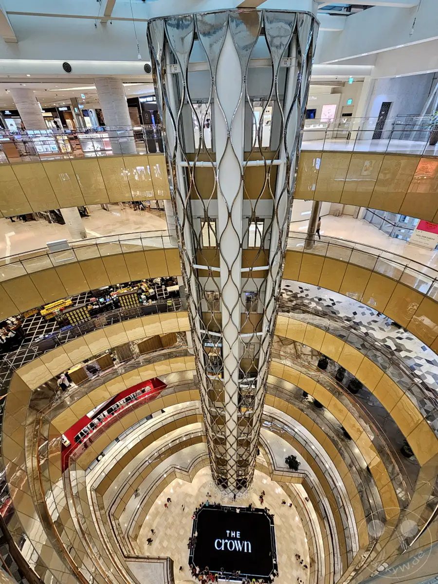 Blick von oben in das Atrium des Lotte World Tower Einkaufszentrums in Seoul, Südkorea. Die geschwungenen Ebenen und der beeindruckende Kronleuchter schaffen eine luxuriöse Atmosphäre. Im Erdgeschoss findet eine Veranstaltung vor einer Leinwand mit der Aufschrift "The Crown" statt.