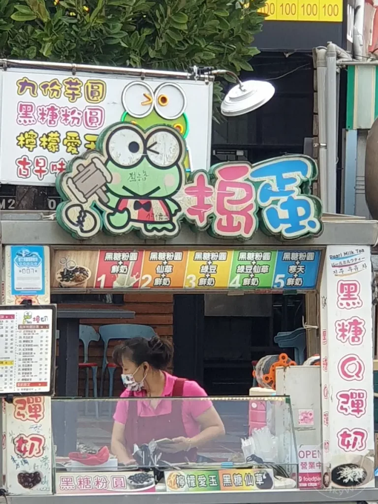 Ein Straßenstand in Taiwan, Kenting, bietet verschiedene Arten von Bubble Tea und Süßspeisen an. Im Vordergrund steht eine Verkäuferin hinter der Theke, während im Hintergrund Speisekarten und bunte Dekorationen zu sehen sind. Die Szene vermittelt eine lebendige und geschäftige Atmosphäre eines lokalen Imbissstandes.