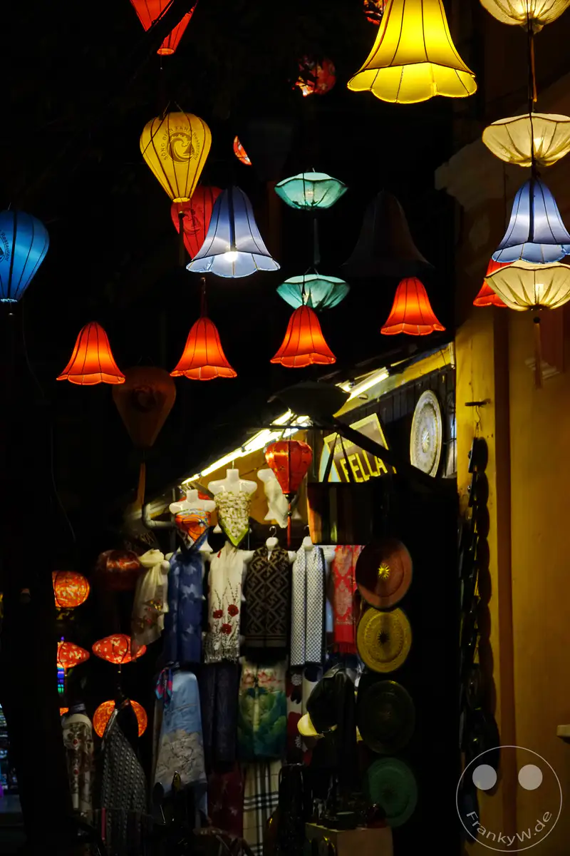 Bunte, beleuchtete Laternen hängen über einer nächtlichen Marktstraße in Hoi An, Vietnam. Textilien und Souvenirs werden in den Läden angeboten, während die atmosphärische Beleuchtung eine magische Stimmung erzeugt. Die Szene strahlt traditionellen Charme und das lebendige Treiben eines südostasiatischen Marktes aus.