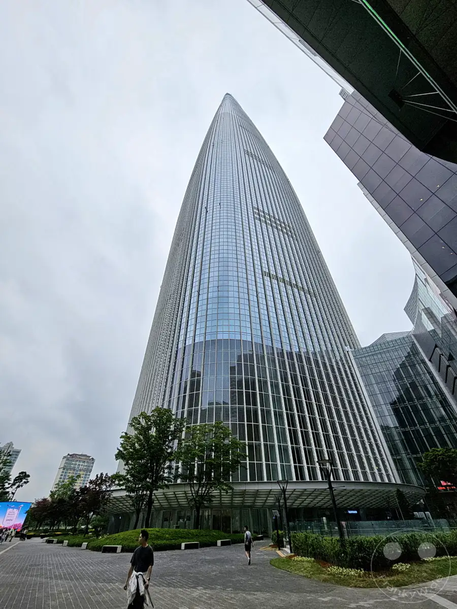 Der Lotte World Tower in Seoul, Südkorea, ragt mit seiner markanten Glasfassade in den bewölkten Himmel. Eine breite Plaza führt zum Eingang des Gebäudes, gesäumt von Bäumen und moderner Bepflanzung. Die Szenerie vermittelt einen Eindruck von urbaner Größe und architektonischer Eleganz.