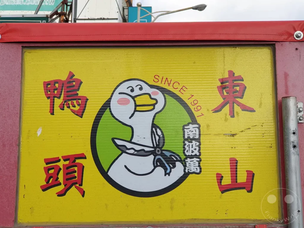 Ein bunter Essensstand in Taiwan, wahrscheinlich in Kenting, wirbt mit einem niedlichen Cartoon-Entenmotiv für sein Angebot. Die gelbe Hintergrundtafel zeigt chinesische Schriftzeichen, die zusammen mit dem Bild eine fröhliche und einladende Atmosphäre schaffen. Die rote Umrandung und die metallischen Elemente des Standes deuten auf eine Outdoor-Verkaufsgelegenheit hin.