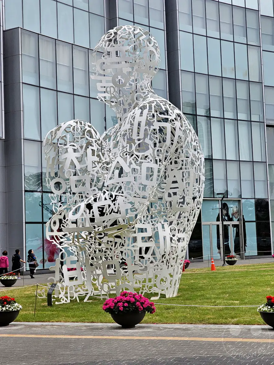 Die beeindruckende Buchstabenskulptur "CommuniT" steht im Vordergrund, mit dem modernen Lotte World Tower in Seoul als Kulisse. Die lebendigen Blumenbeete im Gras verleihen der Szene Farbe und eine einladende Atmosphäre. Menschen gehen im Hintergrund an der Skulptur vorbei und beleben das urbane Umfeld.