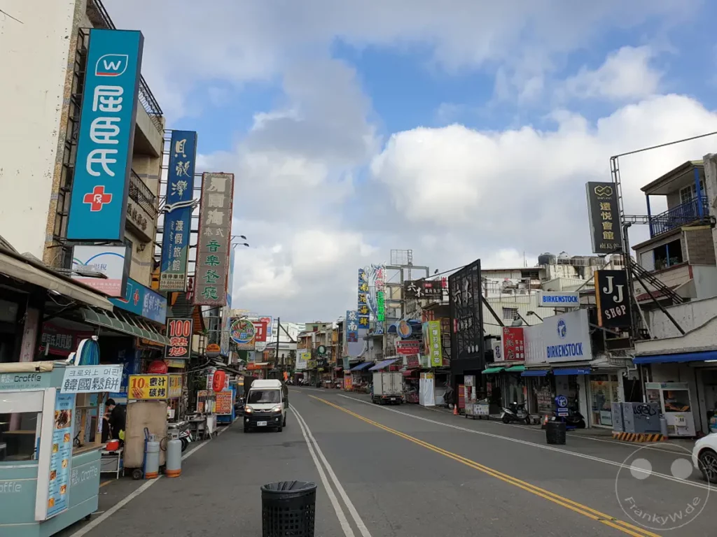 Eine belebte Einkaufsstraße in Kenting, Taiwan, säumt sich mit Geschäften und bunten Schildern, die ein lebhaftes städtisches Ambiente schaffen. Tageslicht wirft Schatten auf den Asphalt, während sich Autos und Fußgänger durch die Szene bewegen. Die Atmosphäre ist geschäftig und typisch für eine touristische Einkaufsgegend.