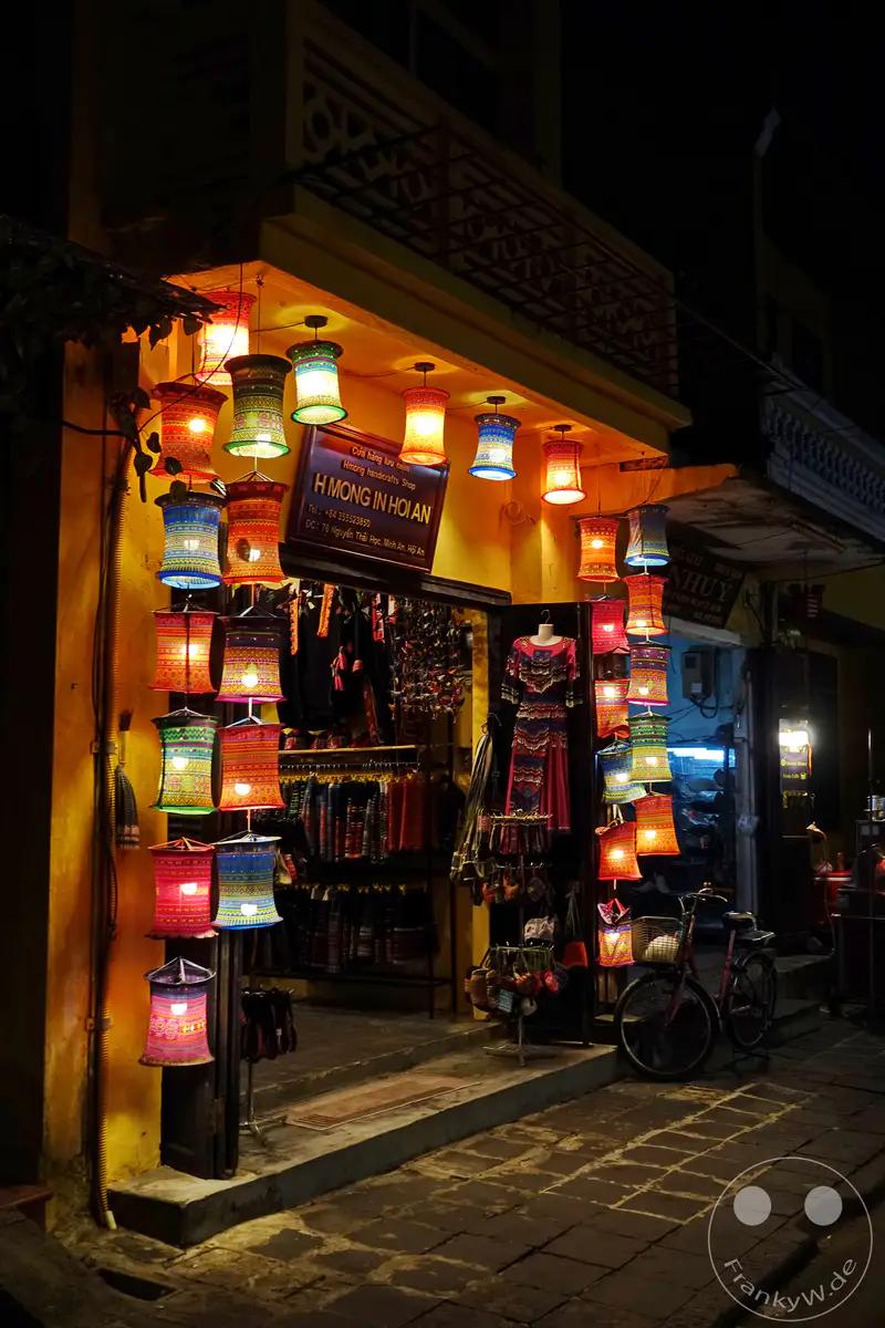 Bunte, leuchtende vietnamesische Laternen hängen vor einem Geschäft für Kunsthandwerk in Hoi An. Nachts wird die Gasse von den farbenfrohen Lichtern erhellt und schafft eine magische Atmosphäre. Die Szene spiegelt die kulturelle Vielfalt und den Charme der historischen Stadt wider.