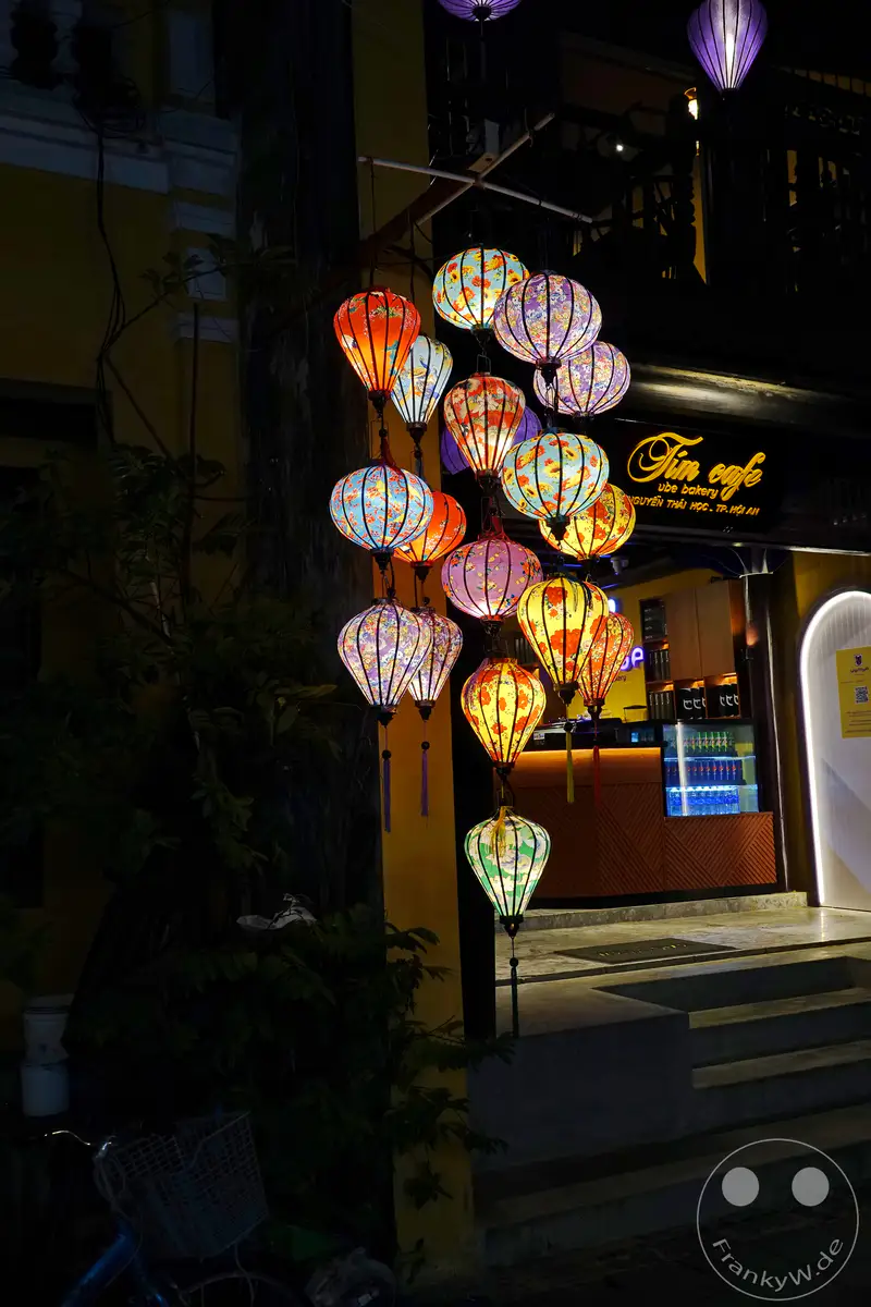 Nachts werden die Straßen von Hoi An mit bunten, traditionellen Laternen beleuchtet, die eine magische Atmosphäre schaffen. Die leuchtenden Ornamente spiegeln sich auf den Fassaden der Gebäude und in den Gassen, während das geschäftige Leben in den Cafés und Geschäften weitergeht. Diese Szenerie strahlt eine festliche und zugleich romantische Stimmung aus, typisch für die Altstadt Vietnams bei Nacht.