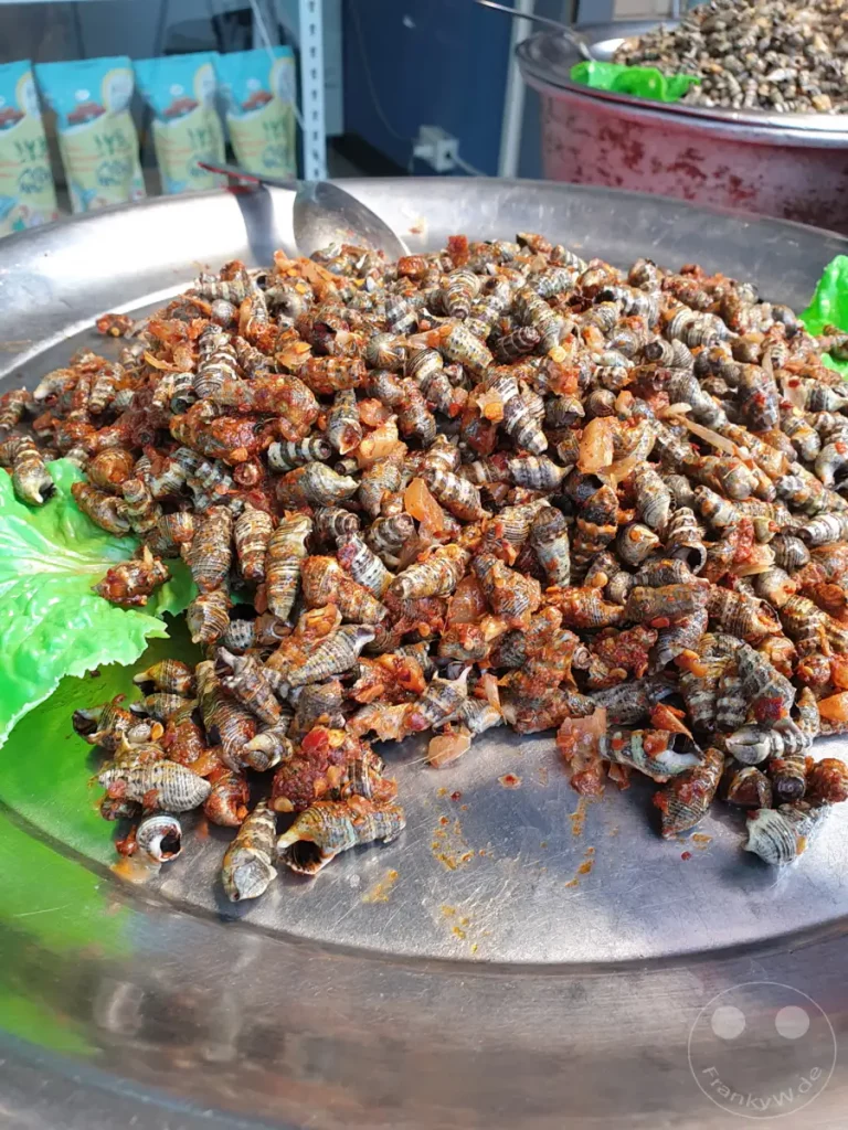 Eine große Portion gewürzter Schnecken liegt auf einem silbernen Blech, garniert mit frischem Salat. Davor eine Schüssel mit weiteren Schnecken im Hintergrund, umgeben von bunten Verpackungen. Die Szene vermittelt den lebhaften Eindruck eines asiatischen Streetfood-Marktes in Taiwan.