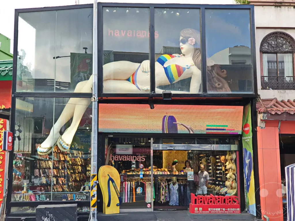 Die Fassade eines Havaianas-Geschäfts in Kenting, Taiwan, präsentiert sich mit einer aufwendigen Dekoration aus einer riesigen Puppe und einer bunten LED-Wand. Im Inneren sind zahlreiche Flip-Flops und Sommerkleidung ausgestellt, die die lebendige Atmosphäre des Ladens unterstreichen. Ein großes gelbes Flip-Flop-Modell steht davor und zieht die Aufmerksamkeit der Passanten auf sich.