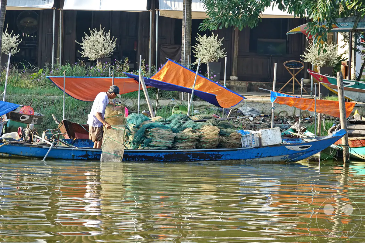 Ein vietnamesischer Fischer sortiert Netze in einem Boot in Hoi An. Die Szene spielt sich auf einem Fluss ab, mit einer Ansammlung von Fischernetzen im Vordergrund und Booten, die am Ufer liegen. Das Wasser reflektiert die umliegende Vegetation und die Gebäude im Hintergrund und vermittelt eine ruhige, alltägliche Atmosphäre des Fischereilebens.