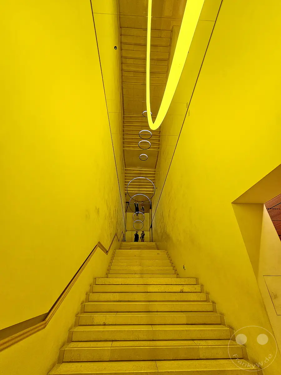 Eine leuchtend gelbe Treppe steigt in einem modernen Gebäude empor, über ihr spiegeln sich futuristische Lichtinstallationen und eine glatte Decke. Die symmetrische Komposition und die intensive Farbe erzeugen eine hypnotische und leicht surreale Atmosphäre im Leeum Museum of Art in Seoul, Südkorea. Architektonische Linien und reflektierende Oberflächen spielen mit der Wahrnehmung und lassen den Raum endlos erscheinen.