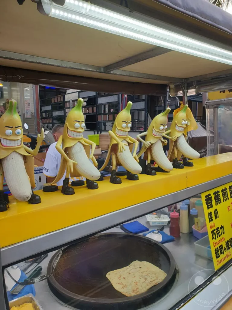 Auf einem Markt in Kenting, Taiwan, stehen mehrere humorvolle Figuren in Form von sprechenden Bananen auf einem gelben Tresen und werben für einen Crepes-Stand. Unter den Figuren wird auf einem heißen Blech gerade ein Crepe zubereitet, was eine lebhafte und unterhaltsame Atmosphäre vermittelt.