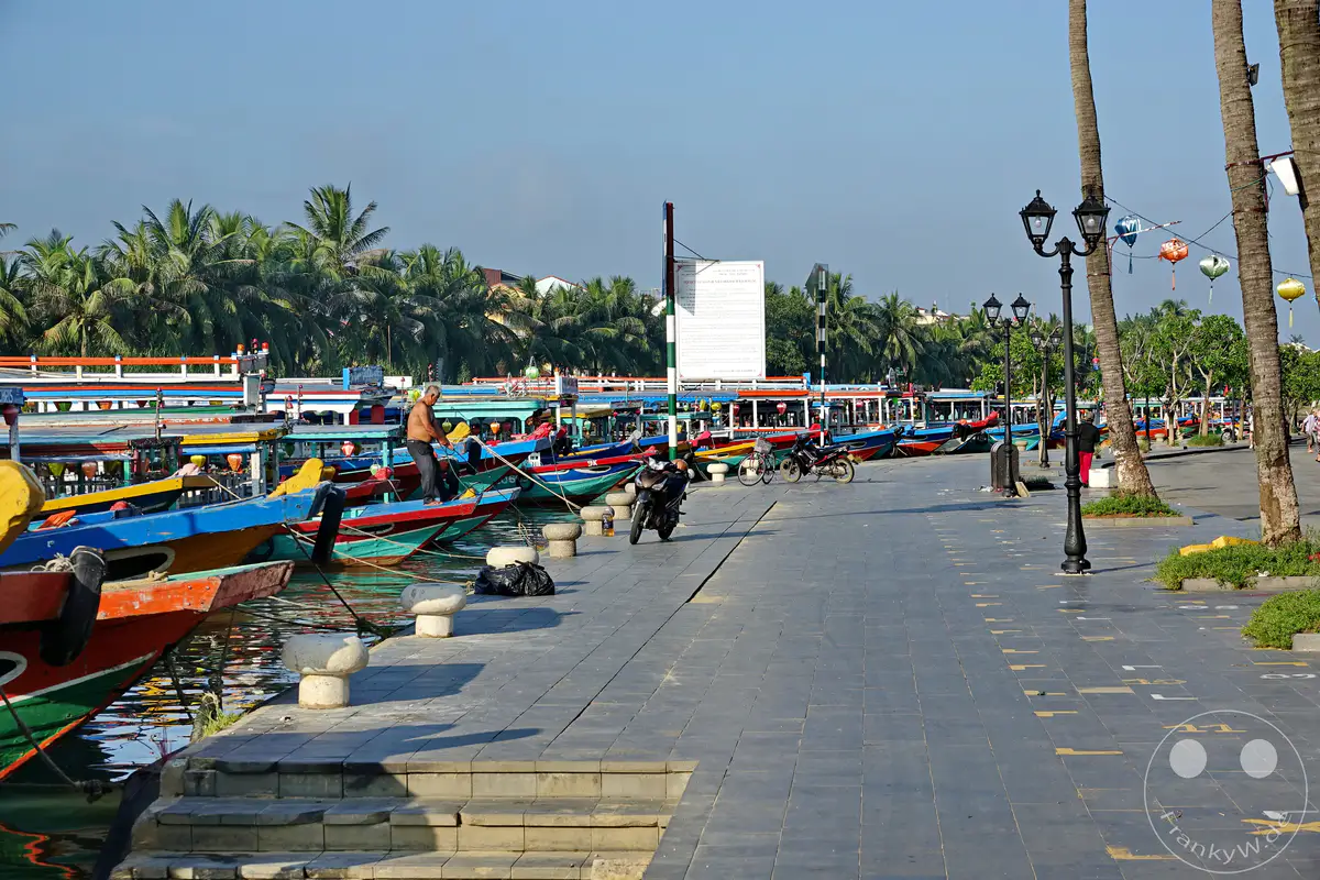 Bunte Ausflugsboote liegen in einer Reihe am Kai des Thu Bon Flusses in Hoi An, Vietnam. Palmen säumen die Promenade, die von Laternen und Laternenpfählen gesäumt wird. Ein Fischer zieht sein Boot am Kai fest, während sich im ruhigen Wasser die bunten Boote und Palmen spiegeln.