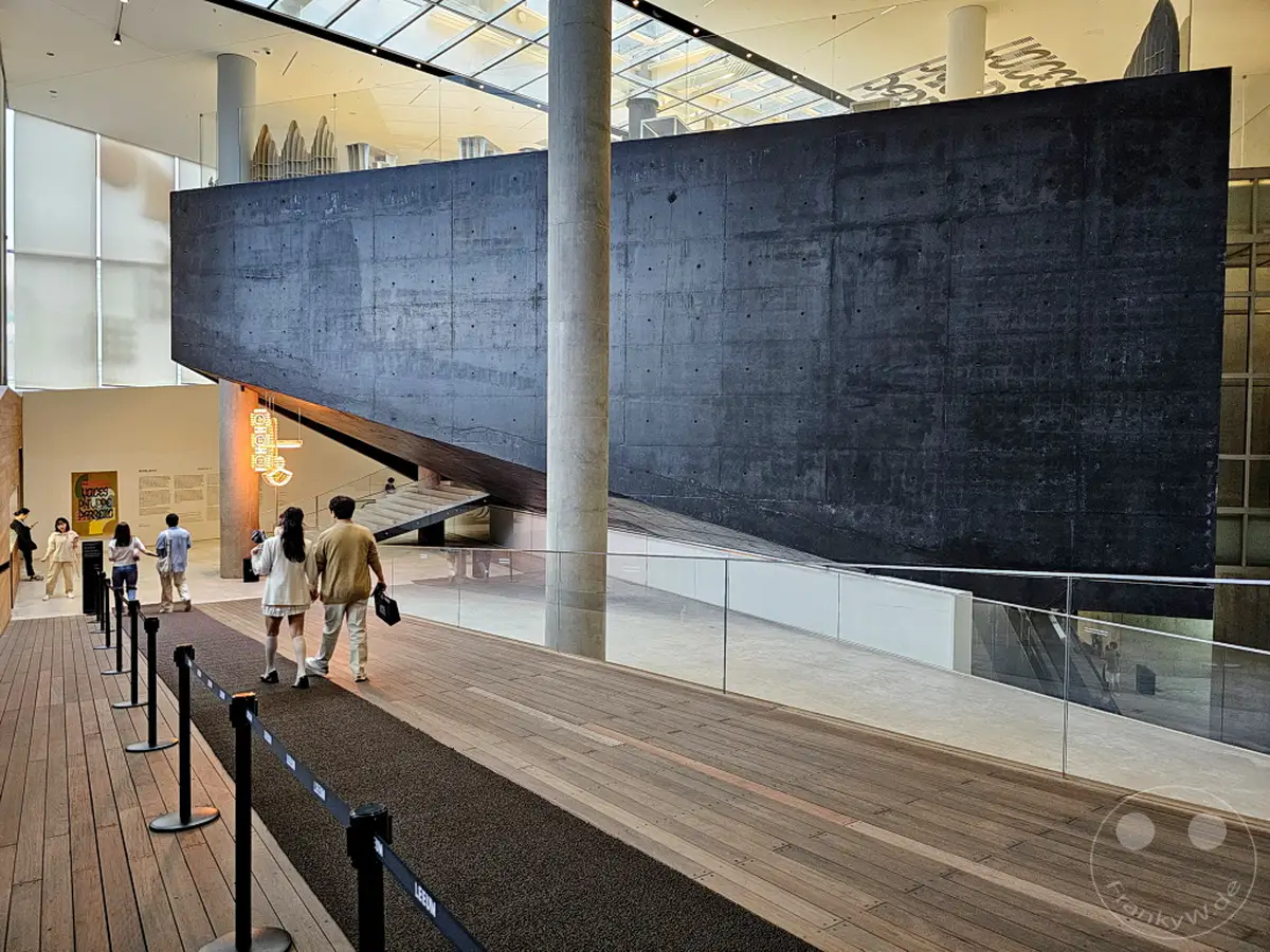 Ein breiter Holzlaufsteg führt durch das Innere des Leeum Museum of Art in Seoul. Eine markante, schräge Betonwand dominiert das Zentrum des Bildes, während Menschen auf dem Gang die futuristische Architektur bestaunen. Das natürliche Licht, das durch die Glaskuppel fällt, erzeugt eine helle und moderne Atmosphäre.