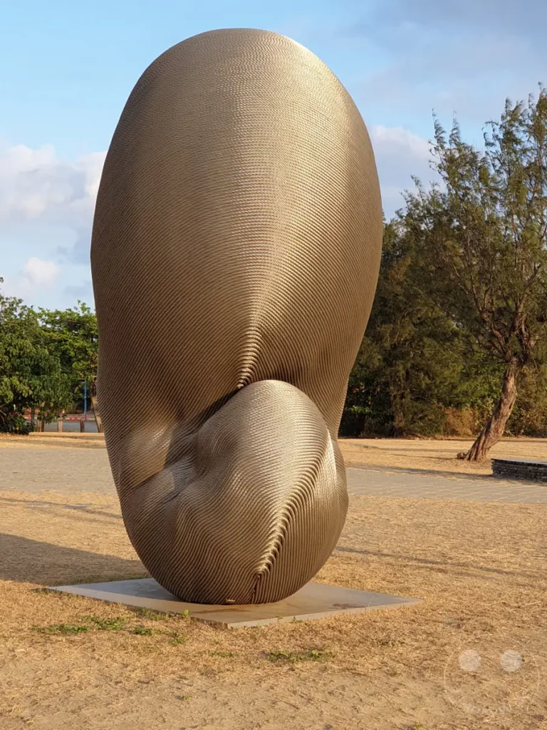 Eine riesige, abstrakte Skulptur aus Metallstreifen steht im Freien in Kenting, Taiwan. Das Kunstwerk "Rebirth" von Kang Muxiang fängt das warme Licht der späten Nachmittagssonne ein und wirft lange Schatten auf das trockene Gras. Die Umgebung ist ruhig und ländlich, mit Bäumen im Hintergrund und einem blauen Himmel mit vereinzelten Wolken, was eine friedliche Atmosphäre schafft.