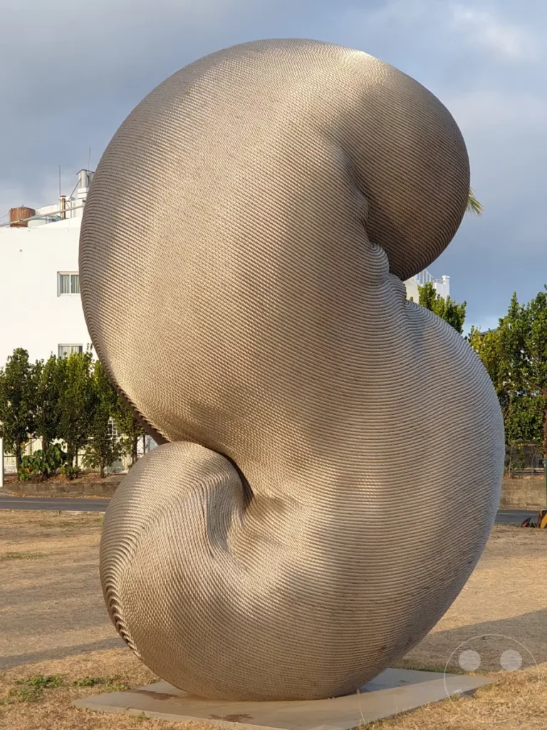 Ein monumentales, spiralförmiges Kunstwerk aus Drahtseilen steht im Freien in Kenting, Taiwan. Die Skulptur "Rebirth" von Kang Muxiang erzeugt durch ihre geschwungene Form und das Spiel von Licht und Schatten eine faszinierende visuelle Dynamik. Die Umgebung mit trockenem Gras und Bäumen im Hintergrund unterstreicht die architektonische Präsenz des Kunstwerks.