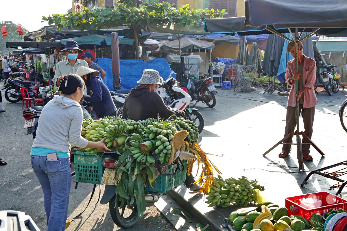 Eine lebhafte Straßenszene auf einem Markt in Hoi An, Vietnam, mit vielen Motorrollern, die für den Transport von Waren, insbesondere von Bananenbündeln, genutzt werden. Die Morgen- oder Abendsonne wirft lange Schatten und beleuchtet die geschäftige Atmosphäre des Marktes. Menschen in traditioneller Kleidung und mit Masken bewegen sich zwischen den Ständen, um die Produkte zu kaufen oder zu verkaufen.